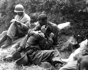 A grief stricken American infantryman being comforted by another soldier. In the background a corpsman methodically fills out casualty tags, Haktong-ni area, Korea. August 28, 1950 